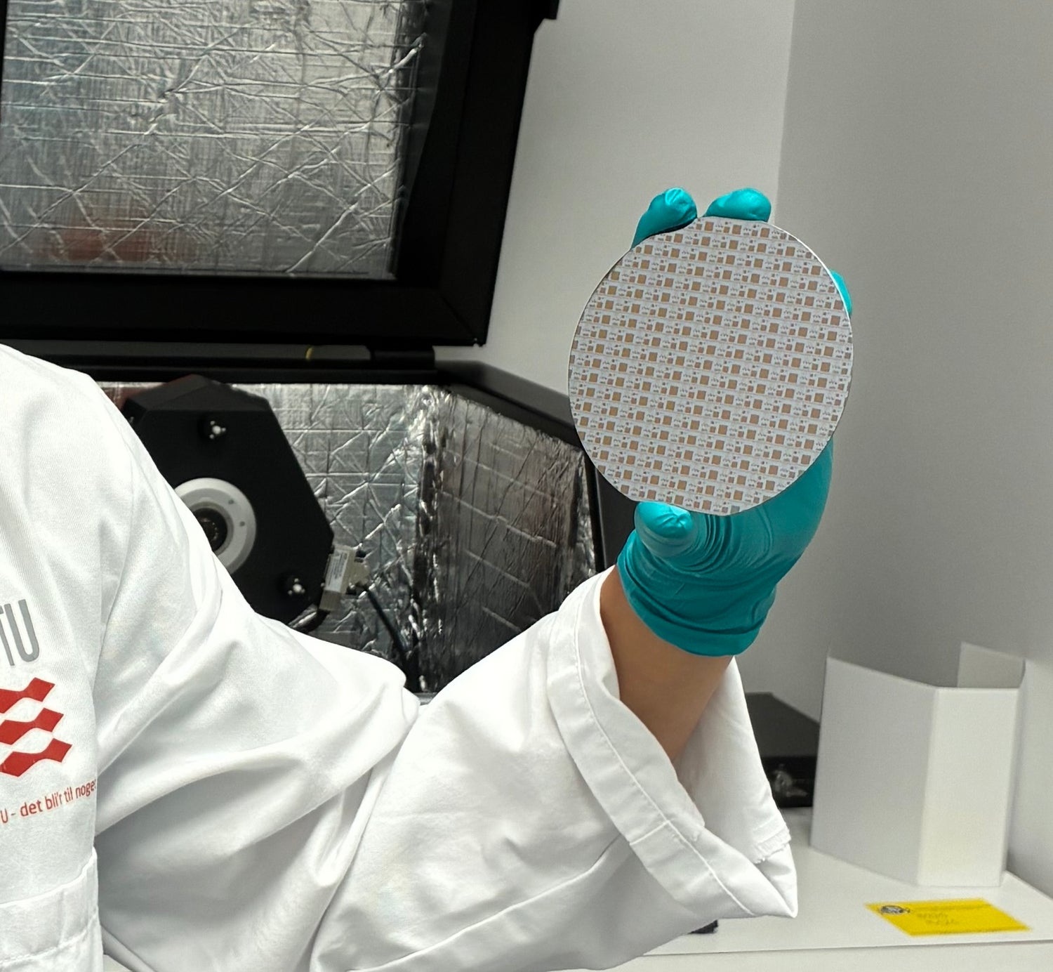 Person in a lab holding a circular wafer with a pattern, wearing a white lab coat and blue gloves.